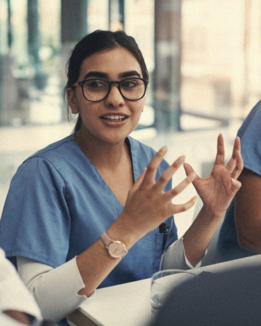A nurse in blue scrubs explaining something to colleagues around a table, mid-sentence, hands raised